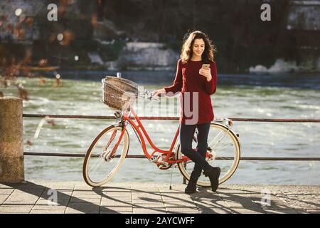 thème du tourisme sur la bicyclette et la technologie moderne. Belle jeune femme caucasienne se tient près de vélo rétro rouge sur la rivière au bord de la rivière Banque D'Images