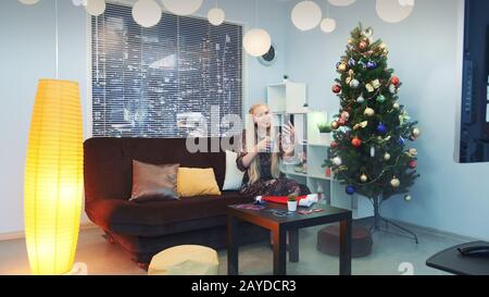 La jeune femme passe des appels vidéo par smartphone avec des gratte-ciel en arrière-plan. Elle est assise sur un canapé avec un verre de boisson dans le salon décoré avec un arbre de noël et des lumières parmi la fumée. Banque D'Images