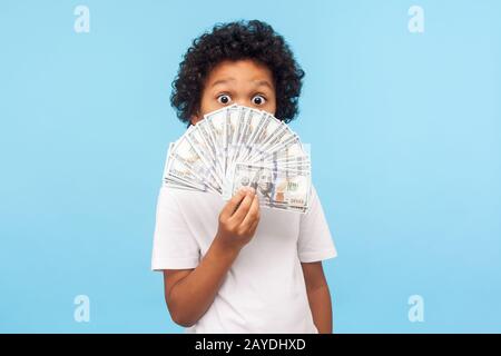 Un joli petit garçon drôle et ébloui avec des cheveux raillés dans un tee-shirt qui cache la moitié du visage avec de l'argent et qui se déchit des billets en dollars avec des yeux stupéfiés. Indo Banque D'Images