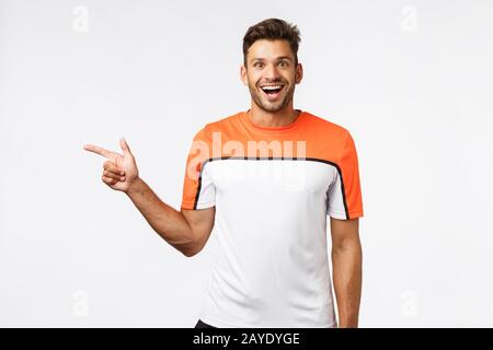 Enchanté de beau sportif barbu dans le t-shirt de sport pointant vers la gauche, voir l'équipement cool dans la salle de sport, se demande et étonné, souriant Banque D'Images