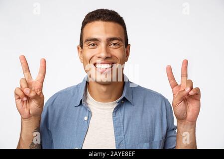 Jeune homme sportif gai dans une tenue décontractée souriant heureux, montrant des signes de paix ou de citation, debout insouciant blanc backgrou Banque D'Images