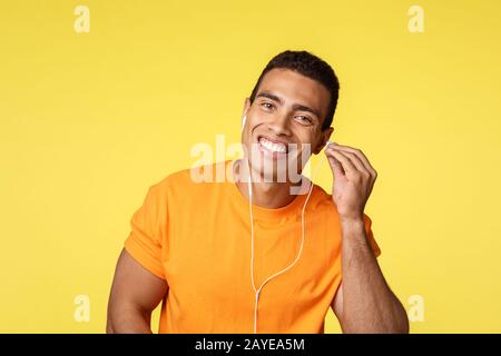 Un bel athlète de sexe masculin dans un t-shirt orange placez un casque dans l'oreille et souriant, sortez, marchez le long de la rue et écoutez le mus Banque D'Images