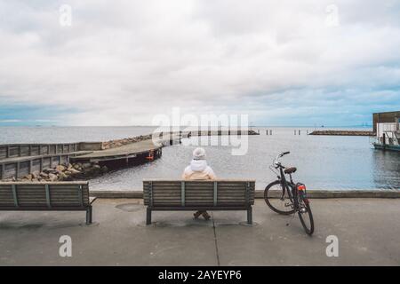 Une jeune femme caucasienne est assise avec son dos sur un banc en bois surplombant la mer Baltique sur le front de mer à Copenhague Danemark in Banque D'Images