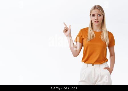 Contrarié, désolé charmante jeune fille blonde moderne dans un t-shirt orange, pantalon, tenir la main dans la poche, pointant vers le coin supérieur gauche, soupirant et sourcils de sillon Banque D'Images