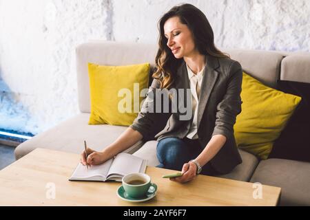 Écrire des produits laitiers sur ordinateur portable dans un café, concept comme mémoire de la vie. Femme avec téléphone mobile et stylo sur table en bois avec tasse de Banque D'Images