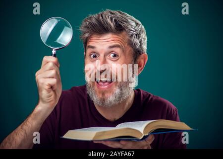 Un portrait d'homme barbu dans un t-shirt rouge tenant un livre ouvert et une loupe. Concept de personnes, d'éducation et de recherche Banque D'Images