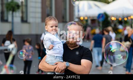 Old Town Square, Riga, Lettonie - 16 août 2019: Homme barbu avec un enfant sur ses mains montres et se réjouit des gigantesques bulles de savon Banque D'Images