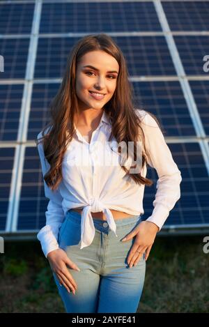 Portrait d'une jeune belle femme souriante avec de longs cheveux bruns debout avec ses mains sur les hanches, module solaire photovoltaïque en arrière-plan Banque D'Images