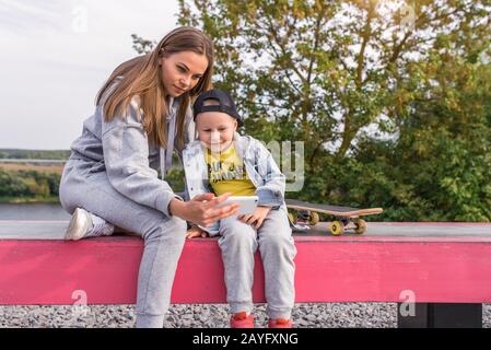 Mère de femme est parent, regarder un film et des photos sur smartphone, Petit garçon enfant de 4-5 ans, skateboard, en été dans la ville sur le terrain de sport. Repos Banque D'Images