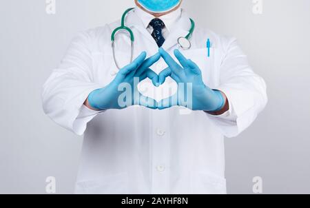 le médecin homme adulte dans un manteau médical blanc avec un stéthoscope sur son cou montre un geste de coeur avec ses mains, portant des gants bleus sur ses mains, blanc Banque D'Images