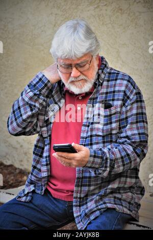 Homme senior assis et essayant de comprendre son vrai nouveau téléphone cellulaire et tous ses changements et avancées avec la barbe et les lunettes en Californie États-Unis Banque D'Images