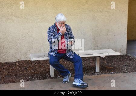 Homme senior assis et essayant de comprendre son vrai nouveau téléphone cellulaire et tous ses changements et avancées avec la barbe et les lunettes en Californie États-Unis Banque D'Images