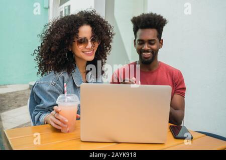 Portrait de deux amis Afro utilisant un ordinateur portable tout en buvant du jus de fruits frais à l'extérieur dans une cafétéria. Banque D'Images