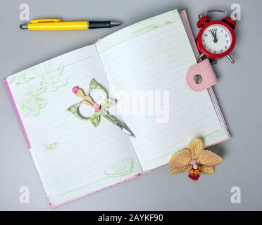 Un carnet ouvert, un stylo, une fleur d'orchidée, des ciseaux et un réveil qui rappelle l'heure sont sur la table. Bureau. Fournitures scolaires. Placer pour le texte. Banque D'Images