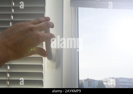 Un jeune homme ferme ou ouvre une fenêtre avec des stores horizontaux en aluminium blanc abaissés. A travers la fenêtre ouverte, les rayons du soleil du printemps du matin se brisent t Banque D'Images