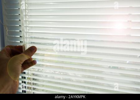 L'homme ferme ou ouvre des stores horizontaux en aluminium blanc abaissés. Par la fenêtre ouverte, les rayons du soleil du printemps du matin se brisent. Banque D'Images