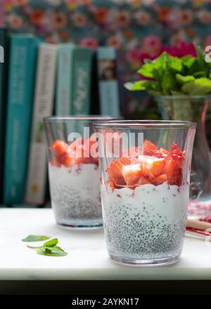 Vue rapprochée de deux verres remplis de morceaux de fraise, de chia, de miel, de coco et de lait. Dans le dos de la scène il y a quelques livres et un pot de Banque D'Images
