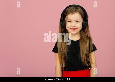 Une petite fille dans un grand casque noir sans fil sur un fond rose pastel regarde l'appareil photo. Le concept de l'écoute et de la musique. Placer pour le texte, l'espace de copie. Banque D'Images