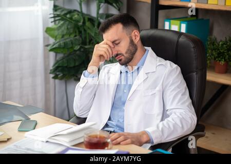 Médecin barbu aux cheveux sombres dans un peignoir blanc qui a l'air fatigué Banque D'Images