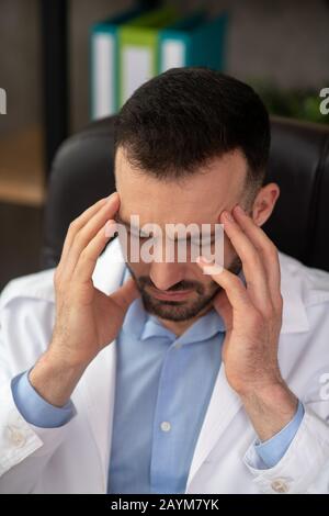 Médecin barbu à poil sombre dans un peignoir blanc ayant mal à la tête Banque D'Images