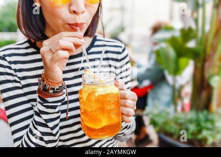 Une heureuse fille hipster élève boit une limonade fraîche à travers une paille dans un café en plein air d'été Banque D'Images