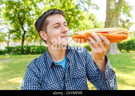 Jeune homme mangeant un grand sandwich de baguette à l'extérieur dans le parc Banque D'Images