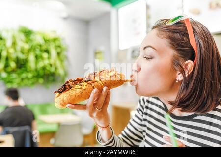 Une jeune fille qui mange un grand hot-dog dans le restaurant fastfood Banque D'Images