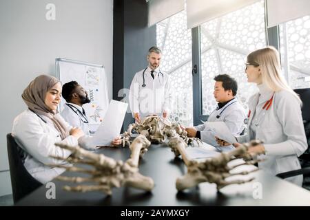 Beau médecin d'âge moyen, professeur d'anatomie, expliquant l'anatomie des os en utilisant le modèle de squelette pour divers élèves en couches blanches. Médecine, éducation Banque D'Images