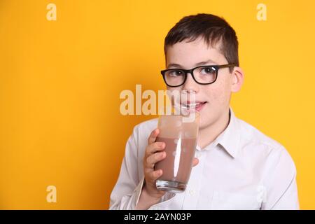 Un jeune garçon de 8 ans boit un verre de lait au chocolat sur fond jaune Banque D'Images