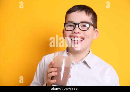 Un jeune garçon de 8 ans boit un verre de lait au chocolat avec une lèvre supérieure recouverte de chocolat sur fond jaune Banque D'Images
