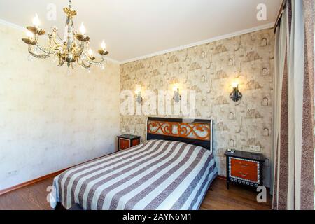 Chambre Classique avec fond d'écran beige. Grand lit avec couvre-lit à rayures noires. Tables de Chevet blanches avec tiroirs rouges. La lampe sur les murs. Banque D'Images