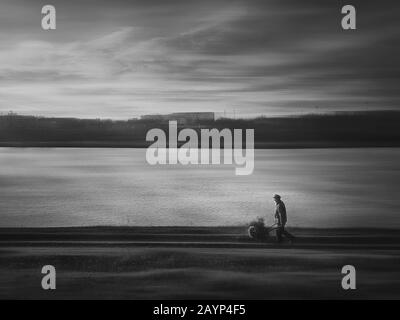 Moody campagne noir et blanc scène, un vieil homme portant un chariot de chariot antique avec bois de chauffage, aller à la maison, marchant une piste de chemin de terre près du lak Banque D'Images