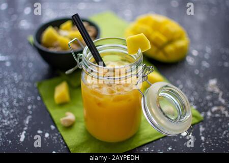 délicieux smoothie à la mangue dans un bol avec paille et glace pilée. Dans le fond flou un muesli avec des fruits et des morceaux de mangue Banque D'Images