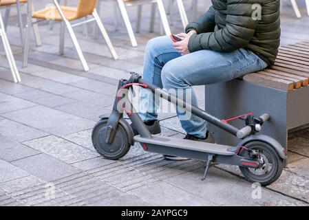 Jeune personne assise sur un toit en regardant son téléphone portable et en se décomposant d'un scooter électrique plié Banque D'Images