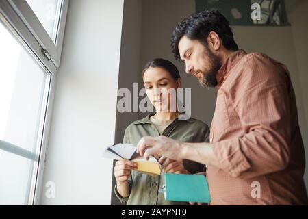 Portrait à faible angle de couple heureux regardant des échantillons de couleur debout par fenêtre tout en redécorant maison, espace de copie Banque D'Images