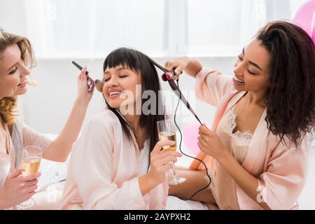 joyeuses amies multiethniques qui font du coiffage, du maquillage et du champagne à la fête de la bachelorette Banque D'Images