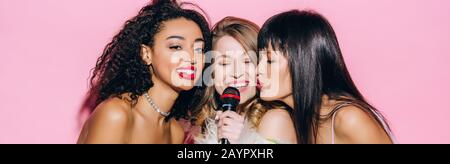 photo panoramique de jeunes filles multiculturelles animées chantant karaoké avec microphone, isolée sur le rose Banque D'Images