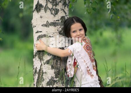 Portrait d'une petite belle fille embrassant un bouleau. Ses épaules sont recouvertes d'un châle aux motifs traditionnels russes. Banque D'Images