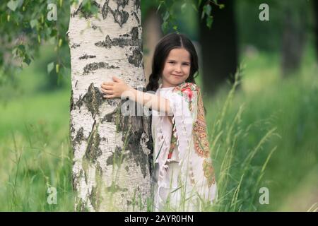 Portrait d'une petite belle fille embrassant un bouleau. Ses épaules sont recouvertes d'un châle aux motifs traditionnels russes. Banque D'Images