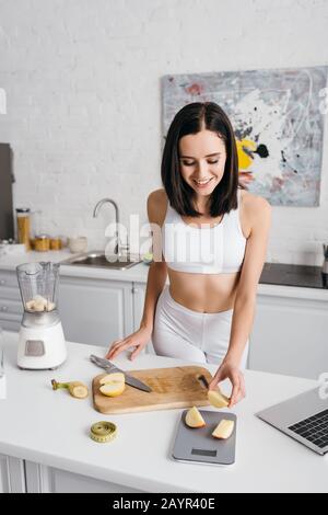 Sportswoman souriant pesant des morceaux de fruits sur les balances tout en préparant le smoothie dans la cuisine, le régime de comptage des calories Banque D'Images