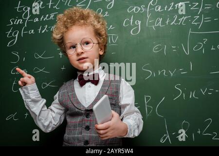 enfant intelligent en costume et noeud papillon tenant le smartphone et pointant avec le doigt près du tableau de surveillance avec des formules mathématiques Banque D'Images