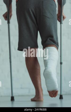 Vue arrière de l'homme avec la jambe dans le bandage en plâtre marchant avec des béquilles Banque D'Images