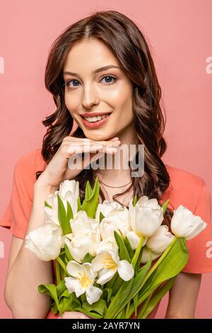 fille rêveuse souriant à l'appareil photo tout en tenant le bouquet de tulipes blanches isolées sur le rose Banque D'Images