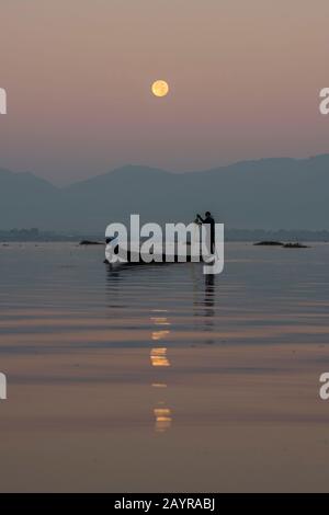 Avant le lever du soleil sous une pleine lune, un pêcheur d'aviron dans son bateau tire dans son filet de pêche il a mis la veille sur le lac Inle au Myanmar. Banque D'Images