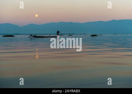 Avant le lever du soleil sous une pleine lune, un pêcheur d'aviron dans son bateau tire dans son filet de pêche il a mis la veille sur le lac Inle au Myanmar. Banque D'Images