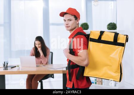 Objectif sélectif de l'homme de livraison avec sac à dos thermo regardant l'appareil photo tandis que femme d'affaires utilisant un ordinateur portable au bureau Banque D'Images