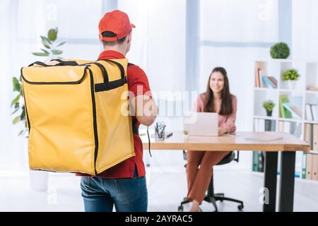 Vue arrière de l'homme de livraison avec sac à dos thermo et femme d'affaires souriante à la table au bureau Banque D'Images