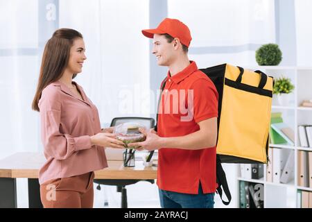 Vue latérale de l'homme de livraison avec sac à dos thermo donnant une salade à emporter à une femme d'affaires souriante au bureau Banque D'Images