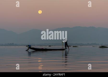Avant le lever du soleil sous une pleine lune, un pêcheur d'aviron dans son bateau tire dans son filet de pêche il a mis la veille sur le lac Inle au Myanmar. Banque D'Images