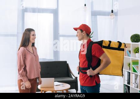 Vue latérale de l'homme de livraison avec sac à dos thermo souriant chez une femme d'affaires au bureau Banque D'Images